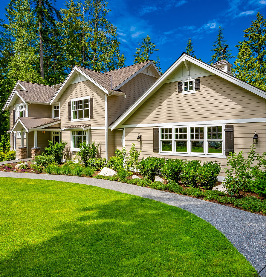 Two-story beige house with white trim and landscaped yard featuring green grass and shrubs.