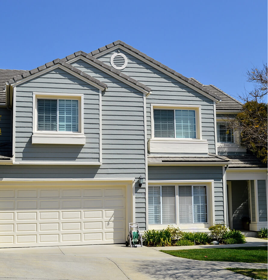 Two-story residential home with gray siding, white trim, and a double garage door, featuring multiple windows and a landscaped front yard.